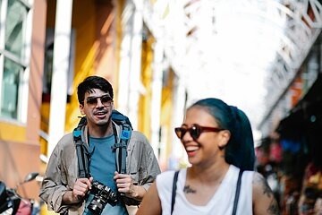 Zwei junge Personen, männlich und weiblich, laufen in Sommerkleidung durch eine Stadt. Die Frau läuft vorn, der Mann lacht sie an, während er eine Kamera in der Hand hält, die um seinen Nacken hängt.
