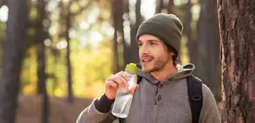 Ein Mann, bekleidet mit einem Hoodie und einer M&uuml;tze, steht in einem Wald. Die Sonne scheint und er setzt eine Wasserflasche zum Trinken an.