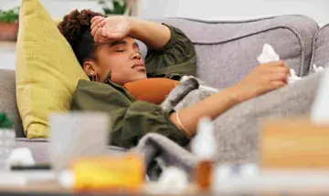 Eine junge Frau liegt auf einem Sofa und h&auml;lt sich ihre Hand an die Stirn. Dabei schaut sie gequ&auml;lt. Im Vordergrund, dabei unscharf, sind Medikamente zu erkennen.