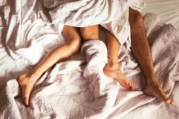 Zwei Personen von denen lediglich die Beine zu sehen sind, liegen halb zugedeckt in einem Bett mit wei&szlig;en Laken. Die Beine sind ineinander verschlungen.