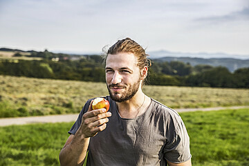 Portrait von Mann, der in einen Apfel gebissen hat. Im Hintergrund grüne Landschaft.