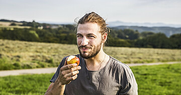 Portrait von Mann, der in einen Apfel gebissen hat. Im Hintergrund grüne Landschaft.