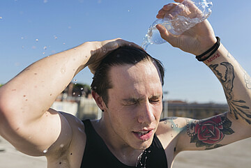 Ein Mann mit Tattoos am Arm schüttet sich Wasser aus einer Falsche über den Kopf.