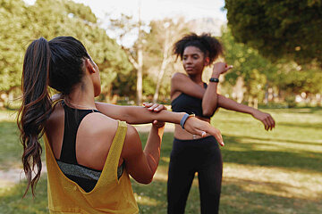 Zwei junge Frauen stehen sich in Sportkleidung in einem Park gegenüber, die Sonne scheint und sie dehnen sich für eine Sporteinheit.