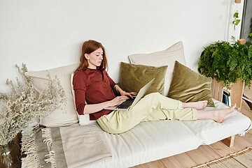 Eine junge Frau sitzt mit Loungekleidung auf einem Sofa, ihre Beine sind ausgestreckt, darauf hat sie einen Laptop abgestellt in den sie schaut.