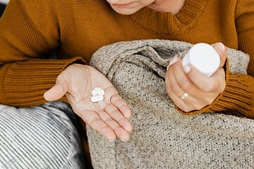 Eine Frau liegt im Bett, zu sehen ist ihre Hand, in der Tabletten liegen. In der anderen Hand hält sie eine Tablettendose.